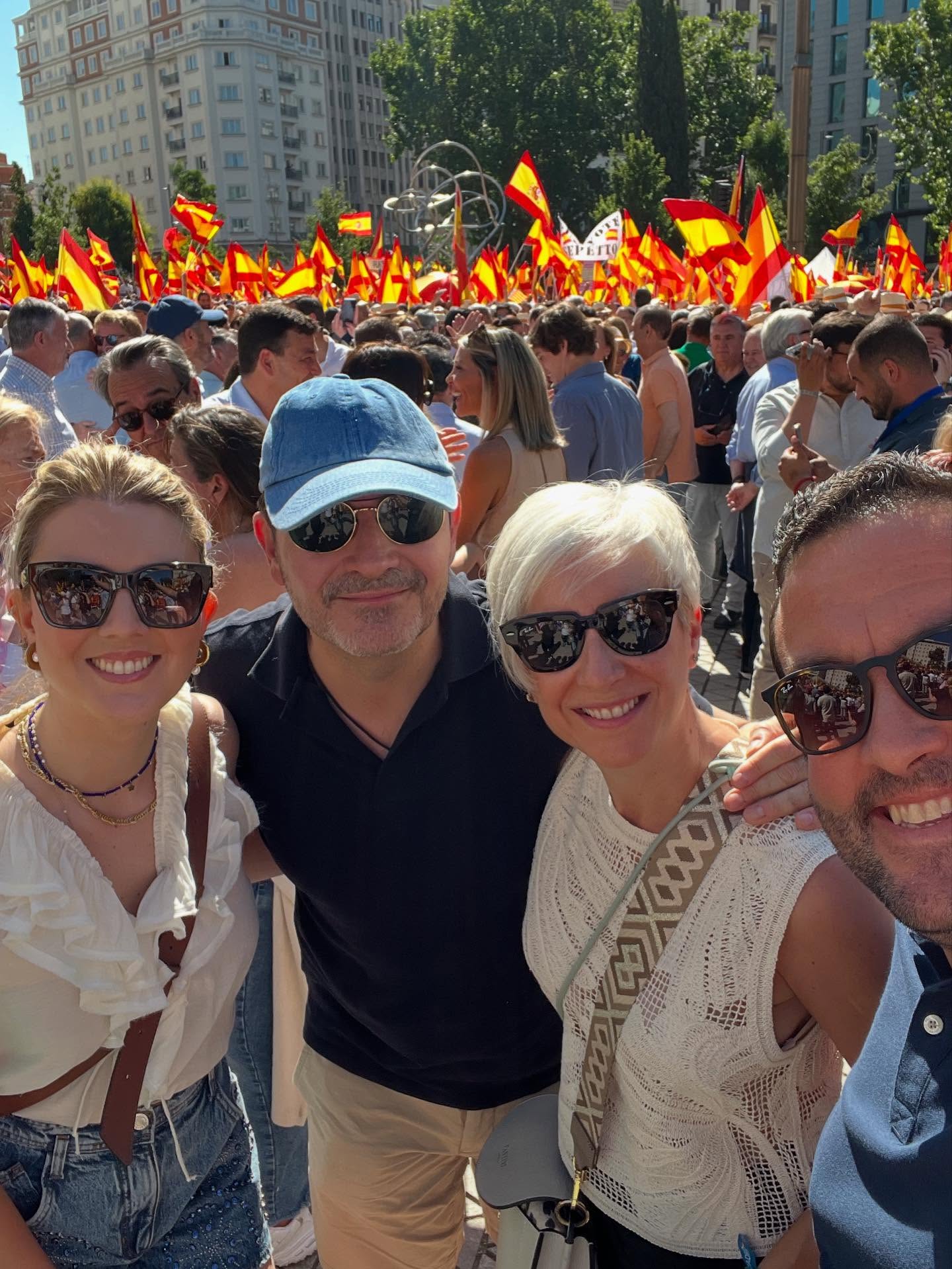 Cuatro personas sonriendo, dos hombres y dos mujeres con gafas de sol, en medio de una concurrida manifestación con banderas de España al fondo.