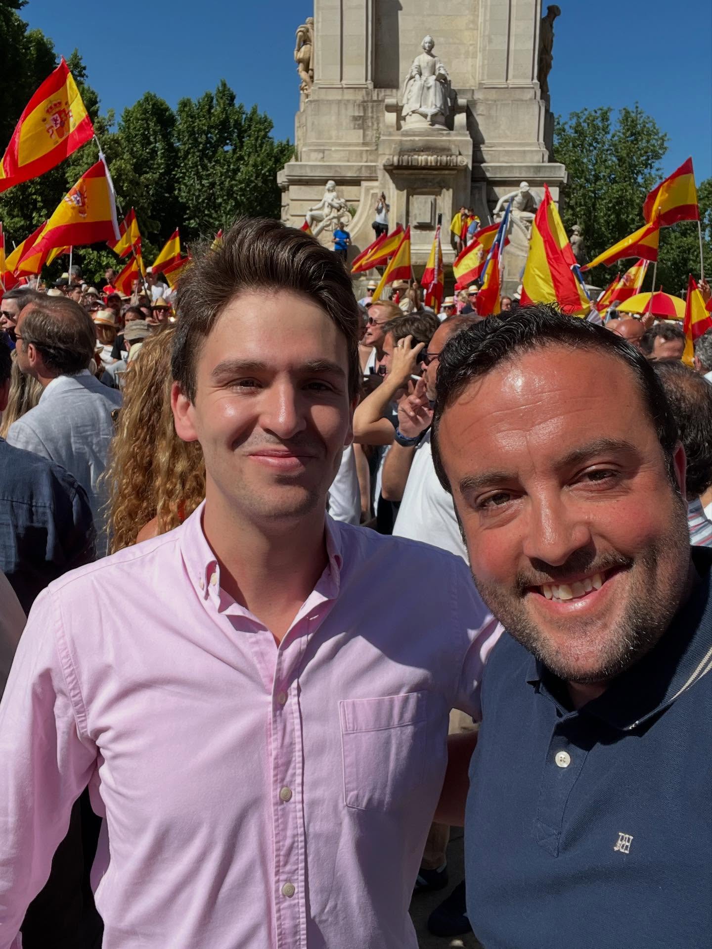 Dos hombres sonriendo en primer plano, con un monumento emblemático y una multitud de banderas de España en una gran manifestación en Madrid.