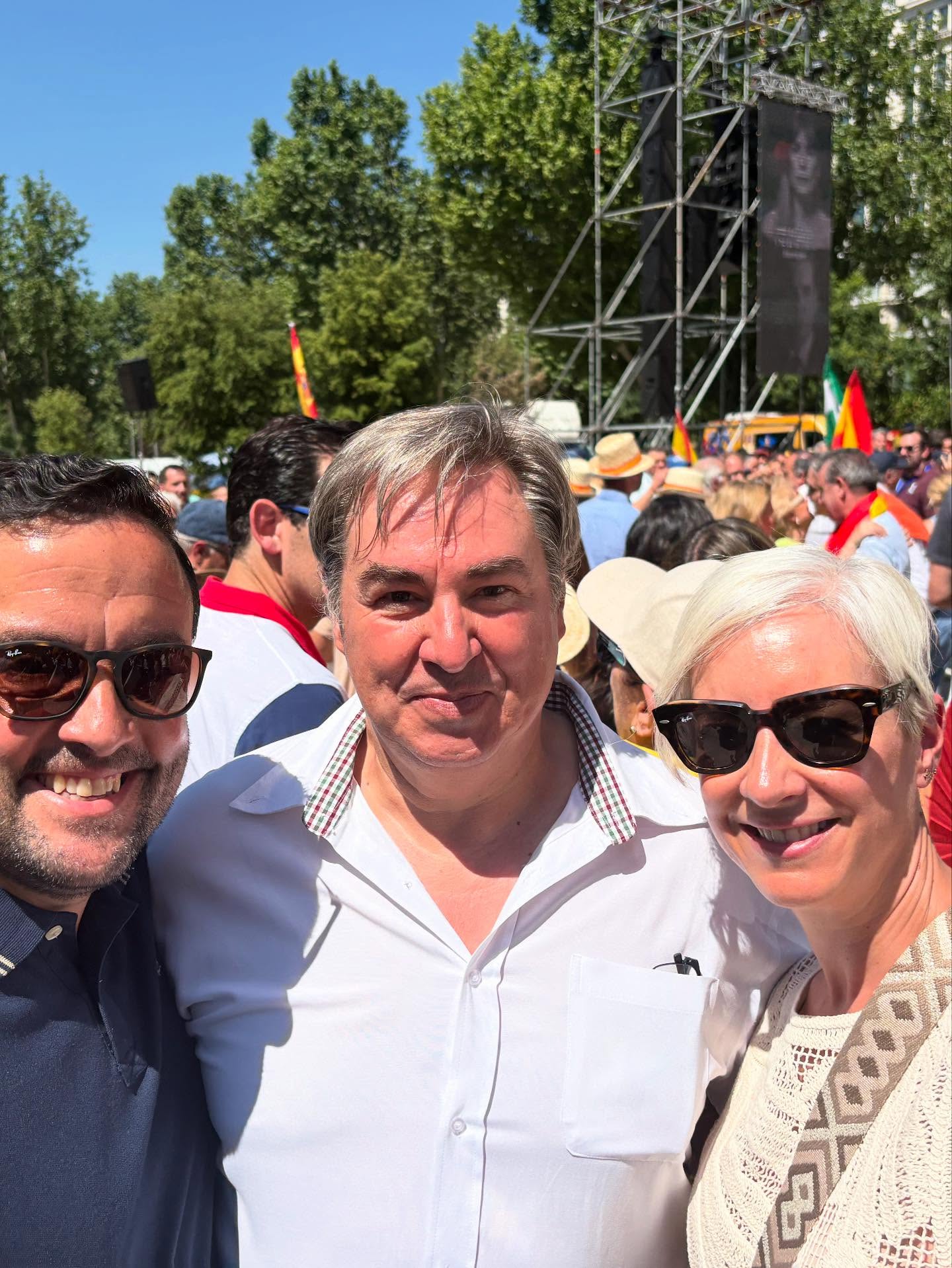 Tres individuos sonrientes, dos hombres y una mujer de pelo corto, posando en primer plano en una concurrida manifestación con banderas de España al fondo.