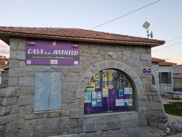Fachada de piedra de la "Casa de la Juventud" con un letrero morado y ventanas cubiertas por contraventanas, en un entorno con árboles y cielo al atardecer.