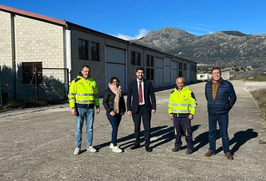El alcalde Sergio Yunquera, de traje, posa con cuatro miembros de su equipo, algunos con chalecos de trabajo, frente a un gran edificio industrial con montañas al fondo.