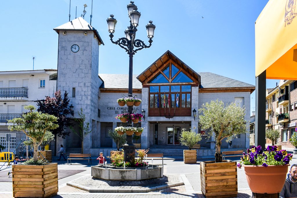 La fachada de la Casa Consistorial de El Boalo con una torre de reloj, una plaza ajardinada con bancos, farolas y fuentes, bajo un cielo azul brillante.