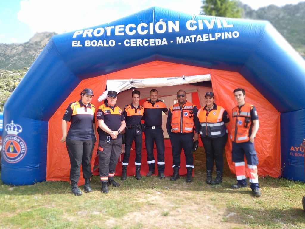 Un grupo de siete miembros de Protección Civil, con uniformes naranjas y azules, posando sonrientes frente a una gran carpa inflable de "Protección Civil El Boalo - Cerceda - Mataelpino".