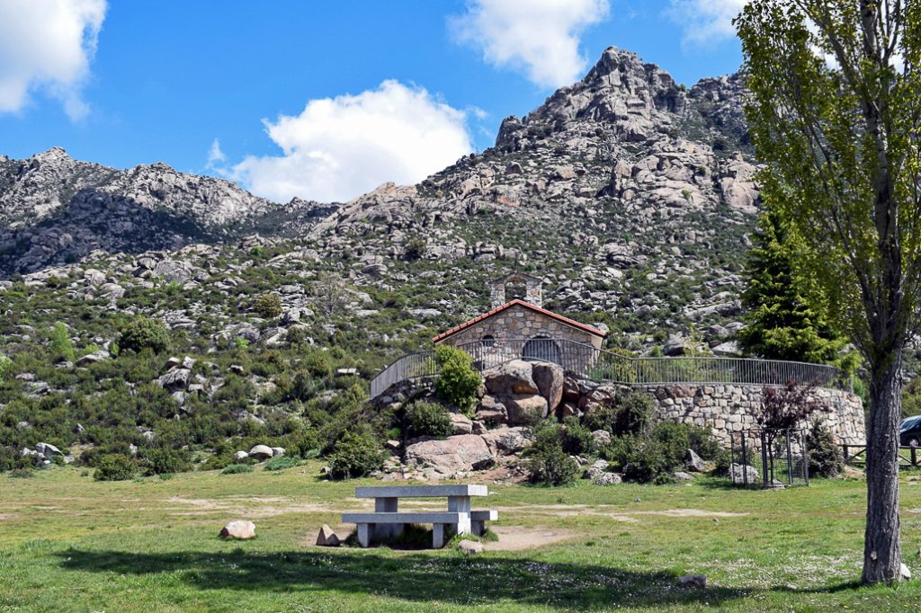 Una pequeña ermita de piedra con tejado rojo, ubicada en una colina rocosa con vegetación, al pie de una gran montaña rocosa, bajo un cielo azul con nubes.