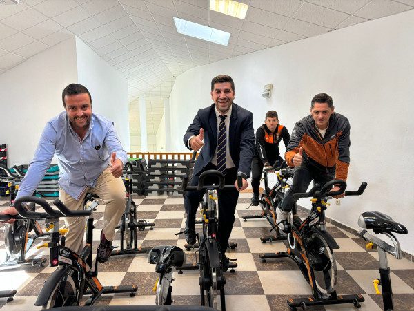 Cuatro hombres sonriendo y haciendo la señal de "pulgar arriba" mientras están sentados en bicicletas estáticas en un gimnasio.
