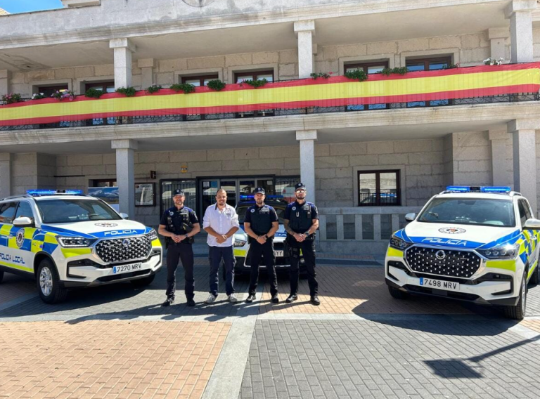 Cuatro hombres, incluido el alcalde Sergio Yunquera y agentes de policía, de pie frente a dos vehículos patrulla nuevos, con la fachada de un edificio adornada con una bandera española al fondo.