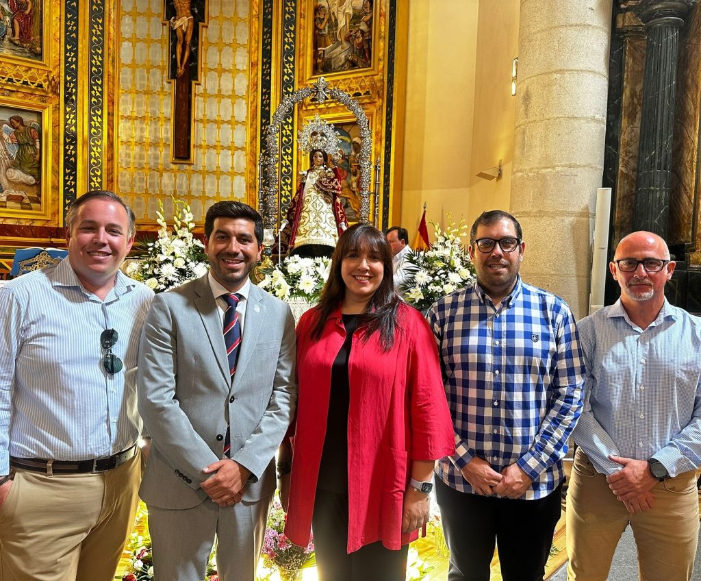 El Alcalde Sergio Yunquera (segundo por la izquierda) y otros representantes, posando sonrientes dentro de una iglesia con la imagen de la Virgen del Espinar decorada con flores en el altar.