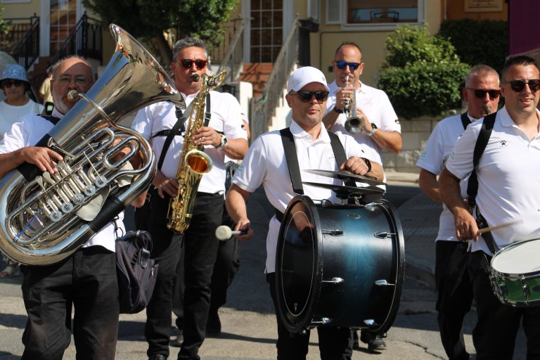 Una banda de música desfilando por las calles de Mataelpino durante las fiestas, con músicos tocando tuba, saxofón y percusión, bajo un cielo azul.
