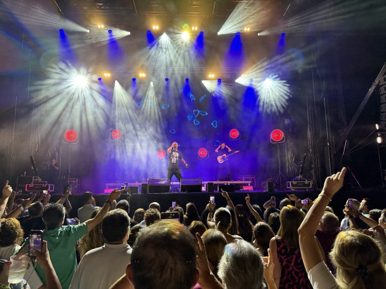 Un concierto nocturno con una banda actuando en un escenario con luces espectaculares, y una multitud de público animando con los brazos en alto.